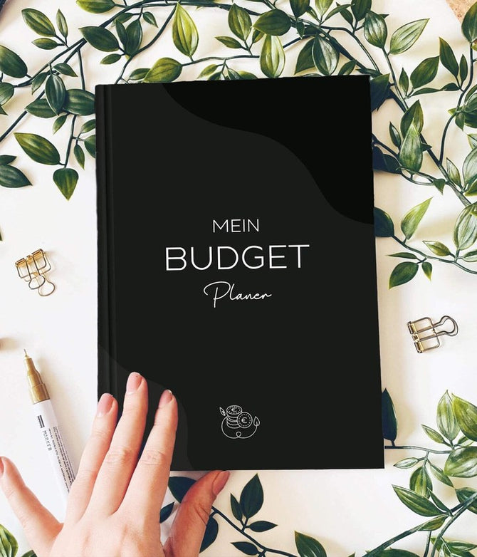 Budgetplaner Schwarz-Weiß von MOMERY in der Hand, umgeben von Blättern